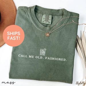 May include: An olive green t-shirt with the text "CALL ME OLD. FASHIONED." and a graphic of a cocktail. The shirt has a crew neck and short sleeves. A gold necklace with a pendant is draped on the shirt.