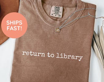 May include: Espresso-colored t-shirt with the text "return to library" in white. The shirt has a crew neck and short sleeves. A silver necklace with a pendant is draped on the shirt. The shirt is folded.