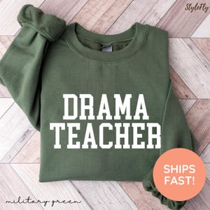 May include: A military green sweatshirt with the words "DRAMA TEACHER" in bold white letters. The sweatshirt has a crew neck and long sleeves. The words "military green" are written in cursive at the bottom of the sweatshirt.