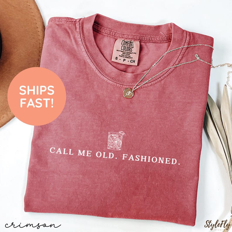 May include: Crimson-colored t-shirt with the text "CALL ME OLD. FASHIONED." and a graphic of a cocktail. A silver necklace with a pendant is draped on the shirt. The shirt has a Comfort Colors label.