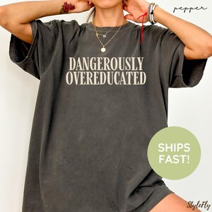 May include: A charcoal gray short-sleeve t-shirt featuring the text DANGEROUSLY OVEREDUCATED in a light beige serif font centered on the chest. The garment has a crew neckline and a relaxed fit with a slightly faded, vintage-inspired appearance.