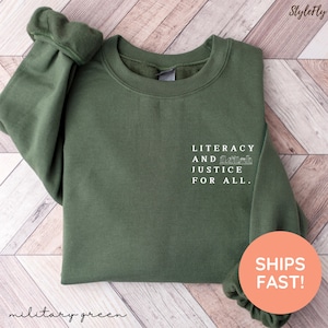 May include: Military green sweatshirt with the text "LITERACY AND JUSTICE FOR ALL." in white. The sweatshirt has a crew neck and long sleeves. The brand name "StyleFly" is at the top right corner.