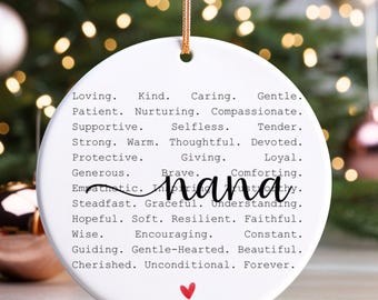 May include: A white ceramic ornament with the word "nana" in cursive and a red heart. The ornament is inscribed with words like "Loving," "Kind," and "Forever." A gold string is attached for hanging. The background is blurred with Christmas lights.