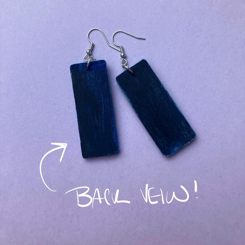 May include: A pair of handmade earrings with a rectangular shape and a dark blue color. The earrings have silver-colored hooks. The text "BACK VIEW!" is written in white on the purple background.