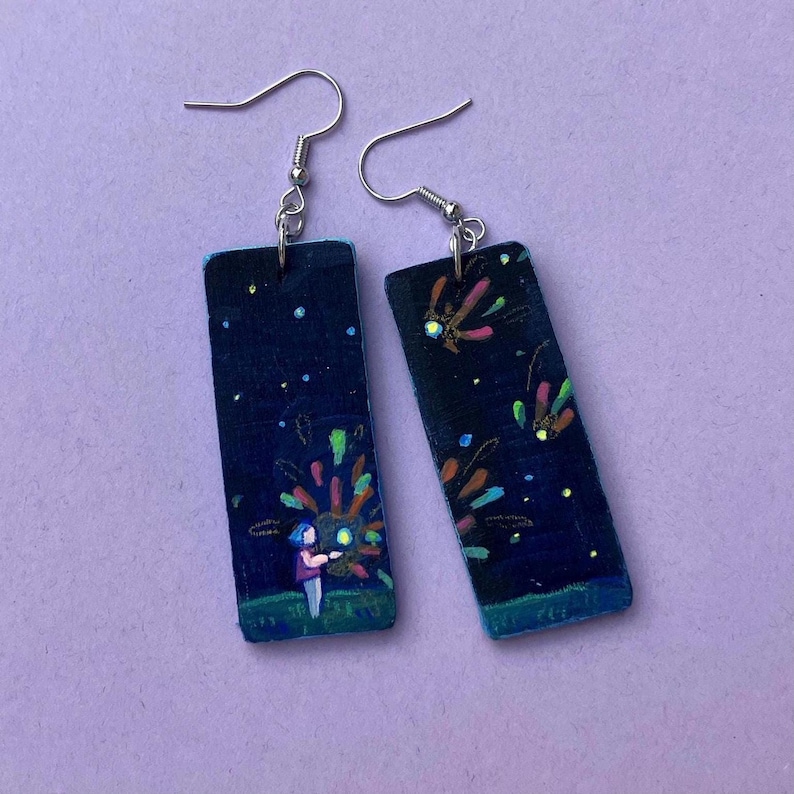 May include: Hand-painted rectangular earrings with a dark blue night sky design. The earrings feature a colorful fireworks display and a small figure. The earrings have silver-colored ear wires.