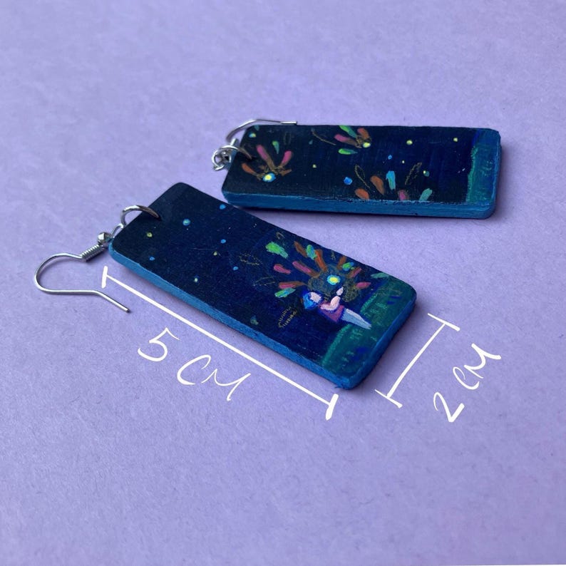 May include: Hand-painted wooden earrings. Each earring is a dark blue rectangle, 2 cm wide and 5 cm long, with a whimsical illustration of a person and fireworks. The earrings have silver-colored ear wires.