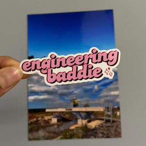 May include: A sticker with the words "engineering baddie" in pink and black, with a small star graphic. The sticker is held in front of a photograph of a construction site with a blue sky.