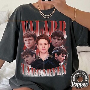 以下が含まれることがあります： 赤毛の男性のグラフィックと「VALARR TARGARYEN」のテキストが赤と白でプリントされた濃い灰色のTシャツ。 シャツはヴィンテージスタイルで、Comfort Colors Pepperのロゴが付いています。