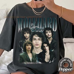 以下が含まれることがあります： フィン・ウルフハードのグラフィックが特徴のダークグレーのTシャツ。「WOLFHARD」という名前が上に、「Finn」という名前が下にあります。 ヴィンテージ感のあるデザインで、俳優の複数の画像が使用されています。「Comfort Colors Pepper」のブランドロゴが見えます。