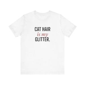 Könnte beinhalten: Weißes T-Shirt mit dem Text "CAT HAIR is my GLITTER" in schwarzer und roter Schrift. Das Shirt ist ein lässiges Kleidungsstück, wahrscheinlich aus Baumwolle oder einem ähnlichen Stoff, geeignet für den Alltag.