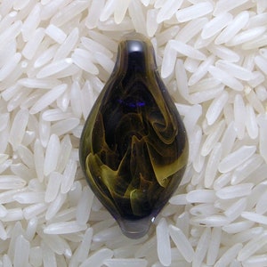 May include: A teardrop-shaped glass pendant with a dark purple exterior and swirling yellow and brown interior patterns. The pendant is set against a background of white rice grains, highlighting its unique design.