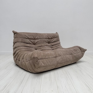 May include: A low-profile, taupe-colored sofa featuring a distinctive tufted, pleated design across the entire surface. The piece has a soft, rounded silhouette with a deep seat and a backrest that curves slightly, creating a continuous, cushioned form.