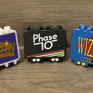 May include: Three colorful card game cases: "Five Crowns" in white and purple, "Phase 10" in black, and "Wizard Card Game" in blue. Each case has a unique design and text, with a rectangular shape and a handle-like top.