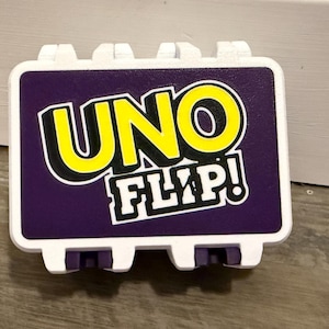 May include: A rectangular white plastic storage case with a purple front panel featuring the yellow and white text UNO FLIP! in a bold, stylized font. The case has a hinged design with visible white tabs along the top and bottom edges.