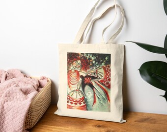 Puede incluir: Un bolso tote beige con una ilustración colorida. La obra de arte representa a una mujer con cabello rojo y un vestido estampado, junto con una figura con una capa negra. El diseño incorpora tonos rojos, blancos y verdes, creando una estética vibrante y artística.