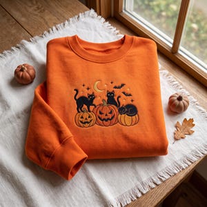 Peut inclure: Un sweat-shirt orange à col rond avec une broderie représentant trois citrouilles et trois chats noirs. Le motif comprend un croissant de lune jaune, de petites étoiles et des chauves-souris en vol, centré sur la poitrine du vêtement.