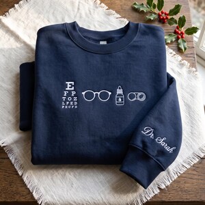 May include: A navy blue crewneck sweatshirt featuring white embroidered graphics on the chest, including an eye chart, eyeglasses, an eye drop bottle, and binoculars. The right sleeve is embroidered with the white cursive text Dr. Sarah.