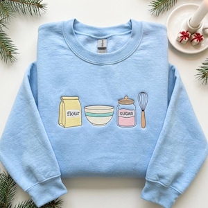 May include: A light blue crewneck sweatshirt featuring an embroidered design of baking essentials. The graphic includes a yellow bag labeled flour, a white mixing bowl with a teal stripe, a glass jar labeled sugar, and a brown handled wire whisk.