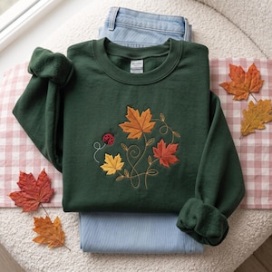 May include: A dark green crewneck sweatshirt featuring an embroidered design of three autumn maple leaves in orange, red, and yellow, accompanied by a small red ladybug and swirling gold stitched vines. The garment has a Gildan brand label.