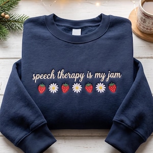 Pode incluir: Uma camisola azul-marinho de gola redonda com o texto bordado "speech therapy is my jam" numa fonte cursiva creme, acompanhada por um padrão horizontal repetitivo de morangos vermelhos e margaridas brancas na zona do peito.