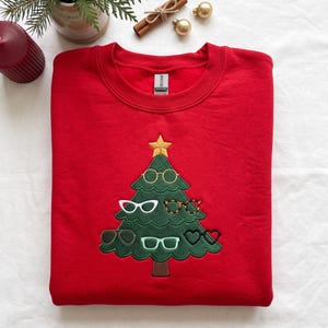 Könnte beinhalten: Ein leuchtend rotes Sweatshirt mit Rundhalsausschnitt und einer gestickten grünen Weihnachtsbaum-Grafik. Der Baum ist mit einem goldenen Stern an der Spitze und sechs verschiedenen Brillen in unterschiedlichen Formen und Farben geschmückt.
