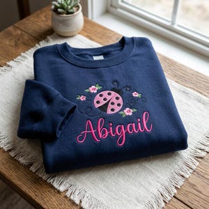 Op de afbeelding: Een marineblauw sweatshirt met ronde hals en een geborduurd ontwerp van een roze lieveheersbeestje met zwarte stippen, omringd door kleine roze bloemetjes en zwarte krullijnen. De naam Abigail is eronder geborduurd in roze cursief schrift.