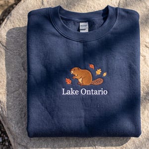 May include: A navy blue crewneck sweatshirt featuring an embroidered brown beaver graphic surrounded by small orange and yellow autumn leaves. Below the beaver, the text Lake Ontario is embroidered in white, sans-serif capital and lowercase letters.