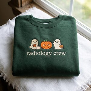 May include: A dark forest green crewneck sweatshirt featuring an embroidered design of two white ghosts and a central orange pumpkin. The ghosts are holding small pumpkins, and the text "radiology crew" is embroidered in white below the figures.