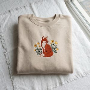 Könnte beinhalten: Ein beigefarbenes Sweatshirt mit Rundhalsausschnitt und einer zentralen Stickerei, die einen rostfarbenen Fuchs zeigt, der zwischen kleinen gelben Blumen mit grünen Stielen und Blättern sitzt. Das Kleidungsstück hat gerippte Bündchen und einen Saum.