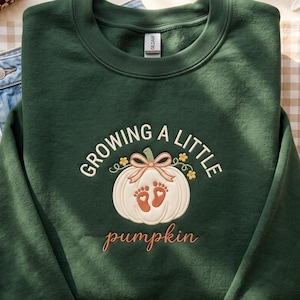 Puede incluir: Una sudadera de cuello redondo verde oscuro con un diseño bordado de una calabaza blanca con un lazo color canela, pequeñas flores amarillas y dos huellas de pies de bebé naranjas. El texto dice "GROWING A LITTLE" y "pumpkin" en cursiva.