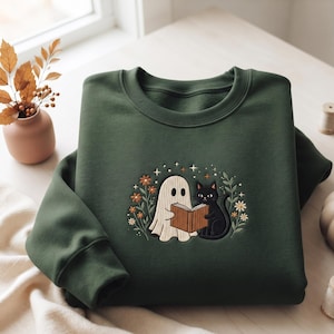 Puede incluir: Una sudadera de cuello redondo en color verde bosque oscuro con un bordado que muestra a un fantasma blanco y un gato negro sentados juntos leyendo un libro marrón abierto, rodeados de pequeñas flores naranjas y blancas y estrellas brillantes.