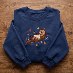Puede incluir: Una sudadera azul marino de cuello redondo con un bordado de una nutria marrón flotando sobre su espalda mientras lee un libro abierto. La nutria está rodeada de pequeñas hojas de otoño de colores en tonos naranja, amarillo y rojo.