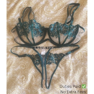May include: A teal lingerie set comprising a bra and thong. The bra features floral lace detailing and adjustable straps. The thong has a delicate design with the text "LOVE YOU" on the waistband. The set is displayed on a patterned surface.