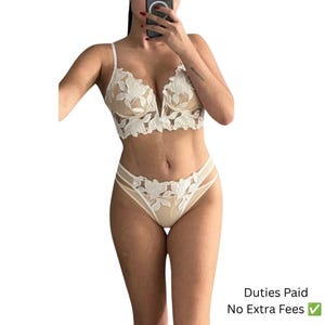 Puede incluir: Un conjunto de lencería de dos piezas que incluye un bralette blanco con bordados florales, escote en V profundo y bragas a juego de corte alto. El conjunto está confeccionado en malla color nude transparente con encaje floral blanco.