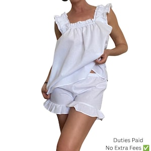 May include: A white two-piece lounge set consisting of a sleeveless top and matching shorts. The top features a square neckline and ruffled shoulder straps. The shorts have an elastic waistband and ruffled hems. The fabric is a solid white color.
