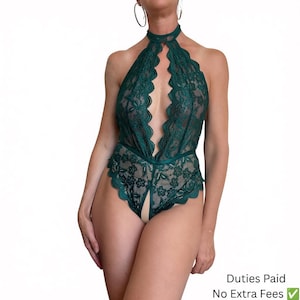 May include: A dark emerald green lace bodysuit featuring a high halter neckline with a choker-style collar, a deep plunging front cutout, and a scalloped lace hemline. The garment has a floral pattern throughout and a vertical opening at the crotch.