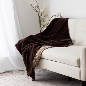 May include: A dark brown throw blanket with a patterned border is draped over a cream-colored sofa. The blanket's texture appears soft and plush, creating a cozy aesthetic. The background includes a white curtain and decorative flowers.