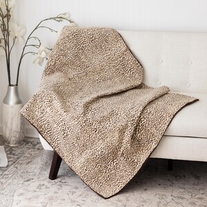 May include: A tan and brown leopard print throw blanket draped over a white sofa. The blanket has a dark brown border and is made of a soft, textured material. The sofa is in a well-lit room with a vase of white flowers.