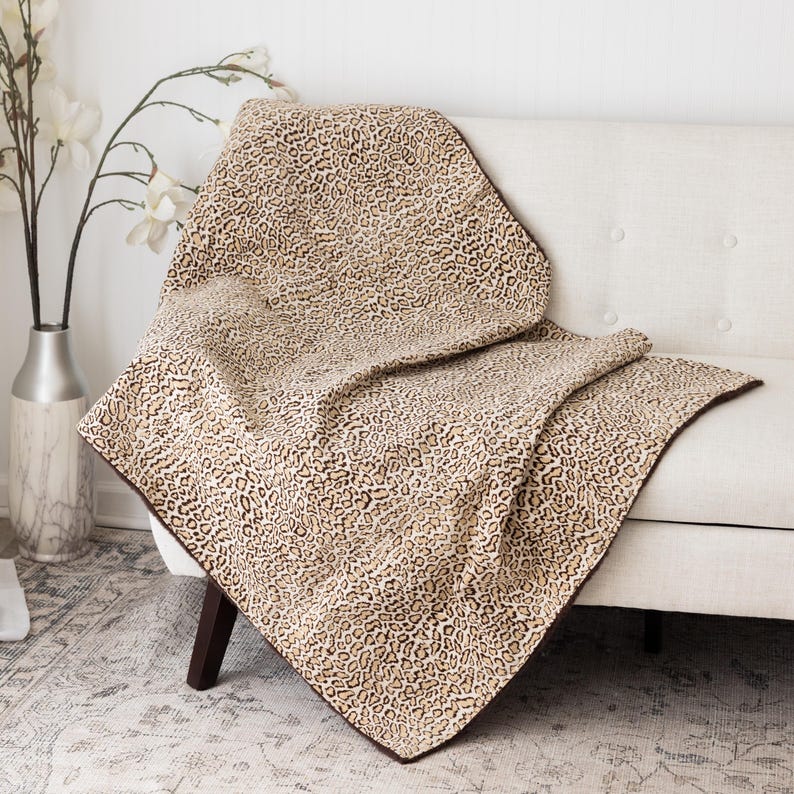 May include: A tan and brown leopard print throw blanket draped over a white sofa. The blanket has a dark brown border and is made of a soft, textured material. The sofa is in a well-lit room with a vase of white flowers.