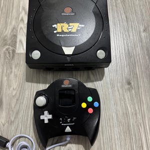 Puede incluir: Consola y mando Dreamcast negros. La consola tiene el logotipo de Dreamcast y el texto "Regulation 7". El mando tiene una cruceta, botones y un cable gris.