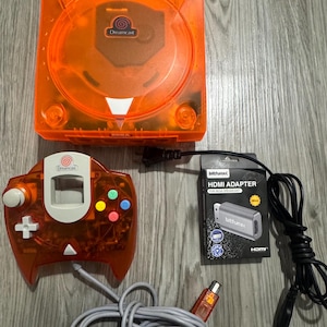 May include: A translucent orange Sega Dreamcast game console with a matching orange controller featuring a white centre panel and colourful buttons. Included is a black Bitfunx HDMI adapter box for Sega Dreamcast with a black power cable attached.