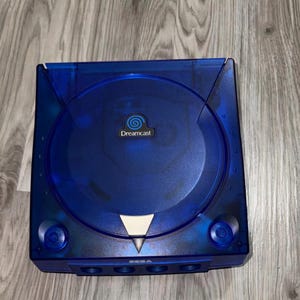 May include: A translucent blue Sega Dreamcast video game console featuring a circular disc lid, a central Dreamcast logo, and a silver triangular accent. The front panel includes four controller ports and power and open buttons.