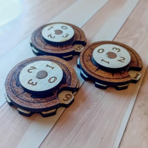 Op de afbeelding: Drie ronde houten speltellers met een donkerbruine, tandwielvormige buitenring en een lichtere beige middenschijf. Elke schijf toont gegraveerde cijfers en een centrale metalen klinknagel, met een kleine uitsparing die één cijfer onthult.