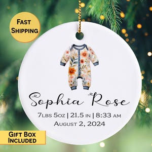 May include: A white ceramic ornament with a floral baby onesie design. The ornament includes the name "Sophia Rose", birth details: 7 lbs 5 oz, 21.5 inches, 8:33 AM, and the date August 2, 2024. The ornament is hanging from a gold string.