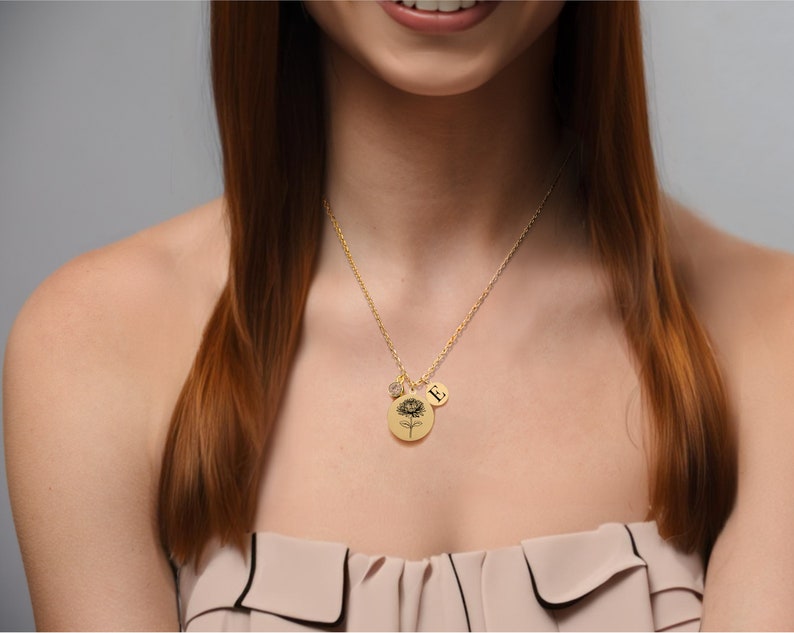 Pu&ograve; includere: Una collana d'oro con una catena delicata e un ciondolo rotondo con un motivo floreale. Il ciondolo ha anche un piccolo ciondolo con la lettera 'E' su di esso.