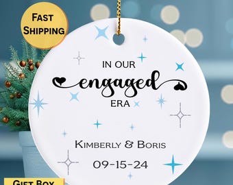 May include: White ceramic ornament with gold hanging cord. The ornament features the text "IN OUR engaged ERA" in black script, with the names "KIMBERLY & BORIS" and the date "09-15-24" below. Light blue star and cross accents decorate the ornament.