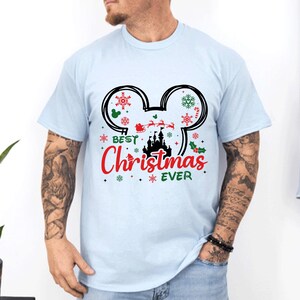May include: A light blue short-sleeve t-shirt featuring a black Mickey Mouse ear outline graphic. Inside the ears are red and green snowflakes, a silhouette of a castle, and Santa in a sleigh. The text reads Best Christmas EVER in red and green.