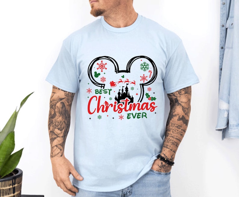 May include: A light blue short-sleeve t-shirt featuring a black Mickey Mouse ear outline graphic. Inside the ears are red and green snowflakes, a silhouette of a castle, and Santa in a sleigh. The text reads Best Christmas EVER in red and green.