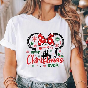 May include: A white short-sleeved t-shirt featuring a graphic of Minnie Mouse ears with a red polka-dot bow, a black castle silhouette, and the text "BEST Christmas EVER" in red and green, surrounded by snowflakes and holiday icons.