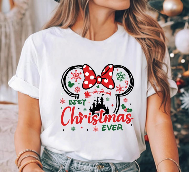 May include: A white short-sleeved t-shirt featuring a graphic of Minnie Mouse ears with a red polka-dot bow, a black castle silhouette, and the text "BEST Christmas EVER" in red and green, surrounded by snowflakes and holiday icons.